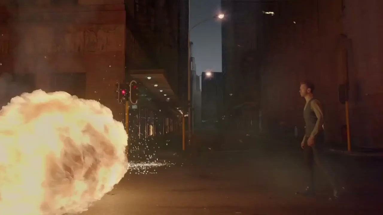 A man in a vest and dark pants walks past a large explosion of fire and sparks in a dark city alleyway at night.