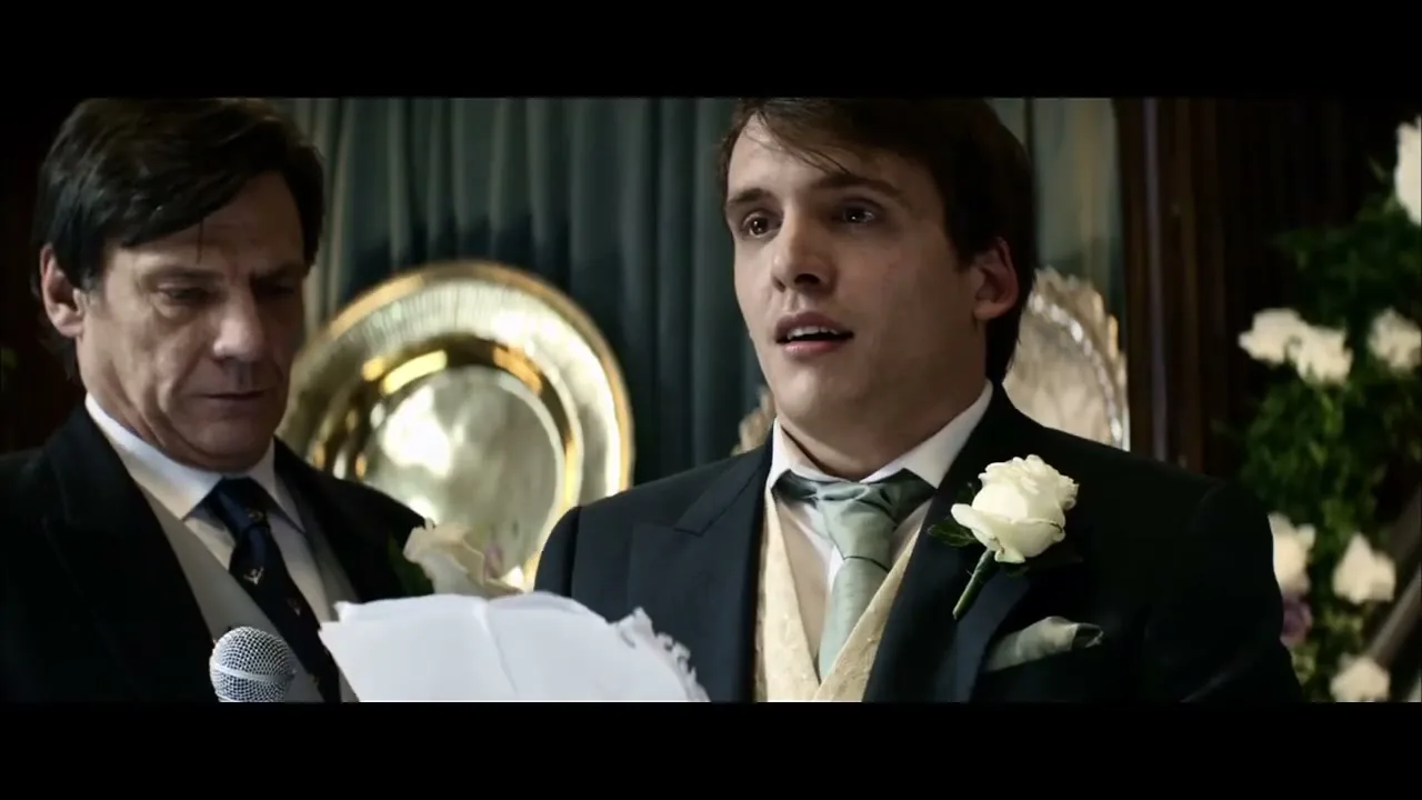 A young man in a suit with a white rose boutonnière looks surprised while holding a microphone and paper, as an older man stands behind him at a wedding reception.