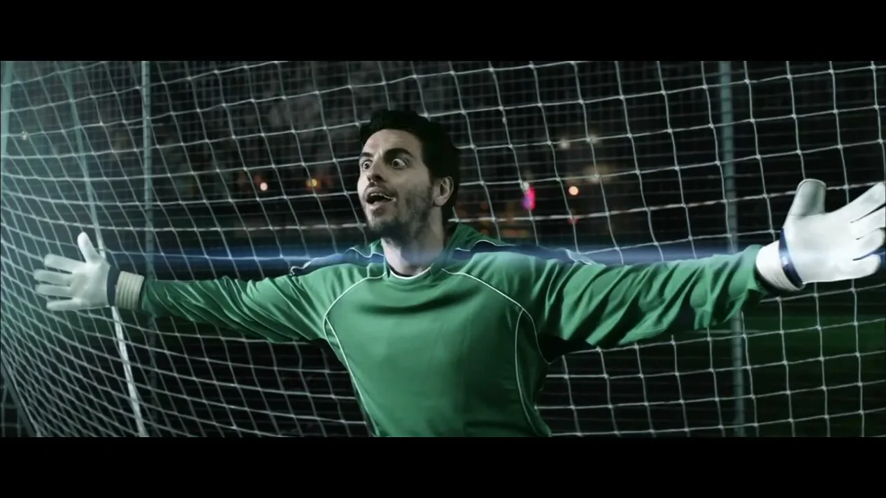 A male soccer goalie in a green jersey and white gloves stands in front of a net, looking up with a surprised expression at night.