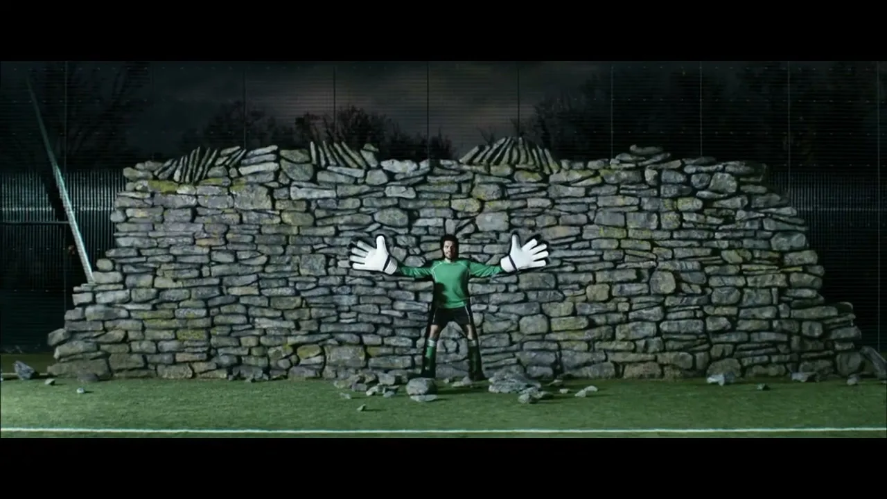 A male soccer goalie in a green jersey and black shorts stands in front of a stone wall, with giant white cartoon hands on his outstretched arms, on a soccer field at night.