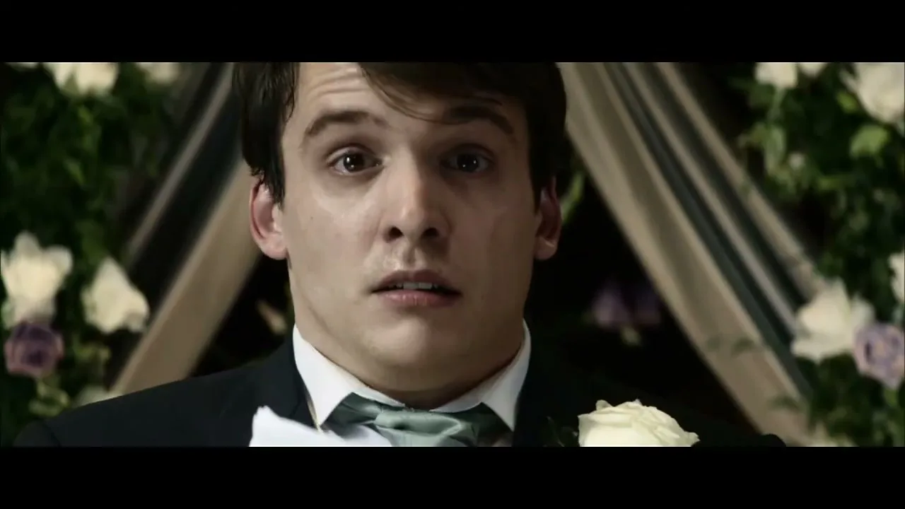 A young man in a tuxedo with a white rose boutonniere looks shocked and nervous, standing in front of a floral archway at a wedding.