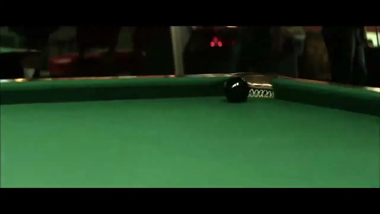 A black pool ball rolls towards a pocket on a green pool table, with a white cue ball blurred in the foreground.