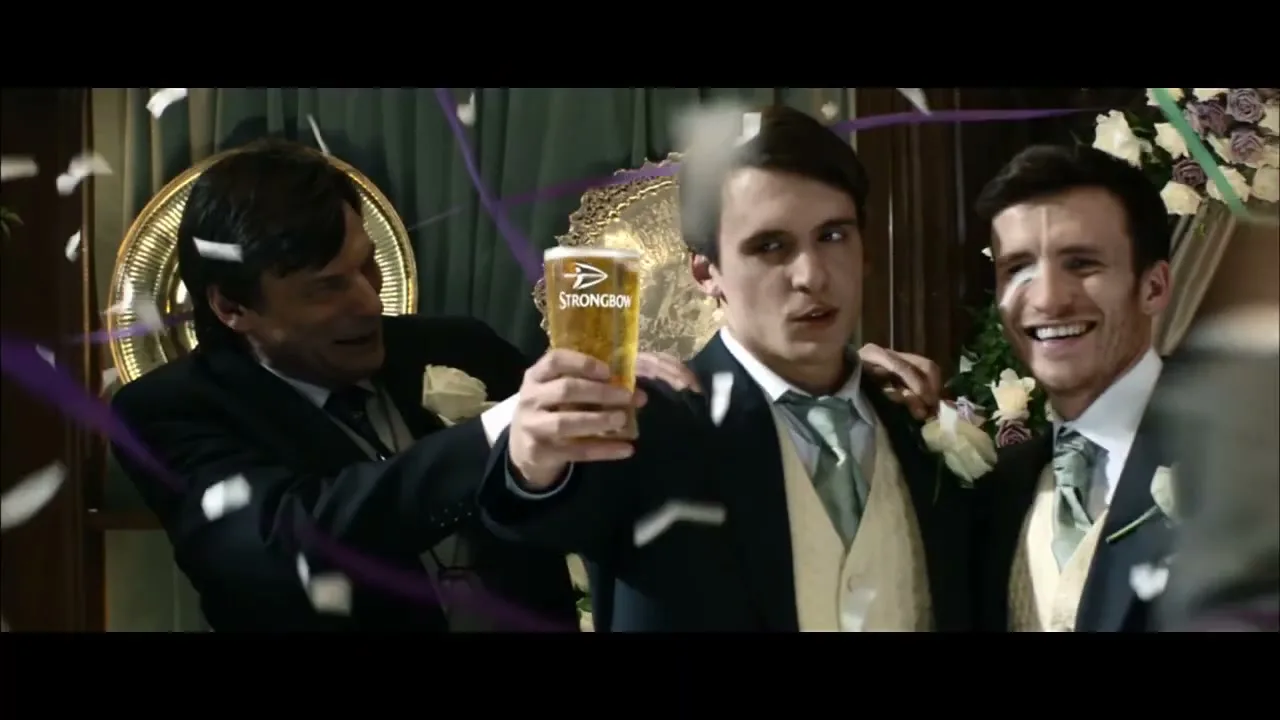 Three men in suits celebrate at a wedding reception, with one man holding up a pint of Strongbow cider as confetti falls around them.