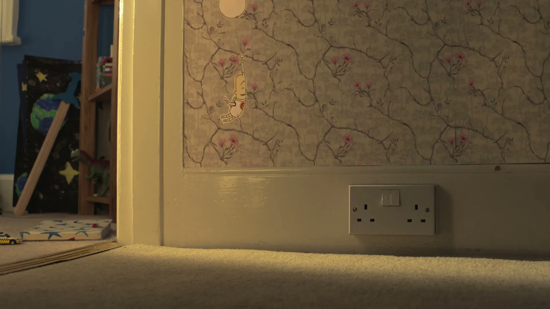 A low-angle shot of a wall with floral wallpaper and a power outlet, with a child's room visible through an open doorway.