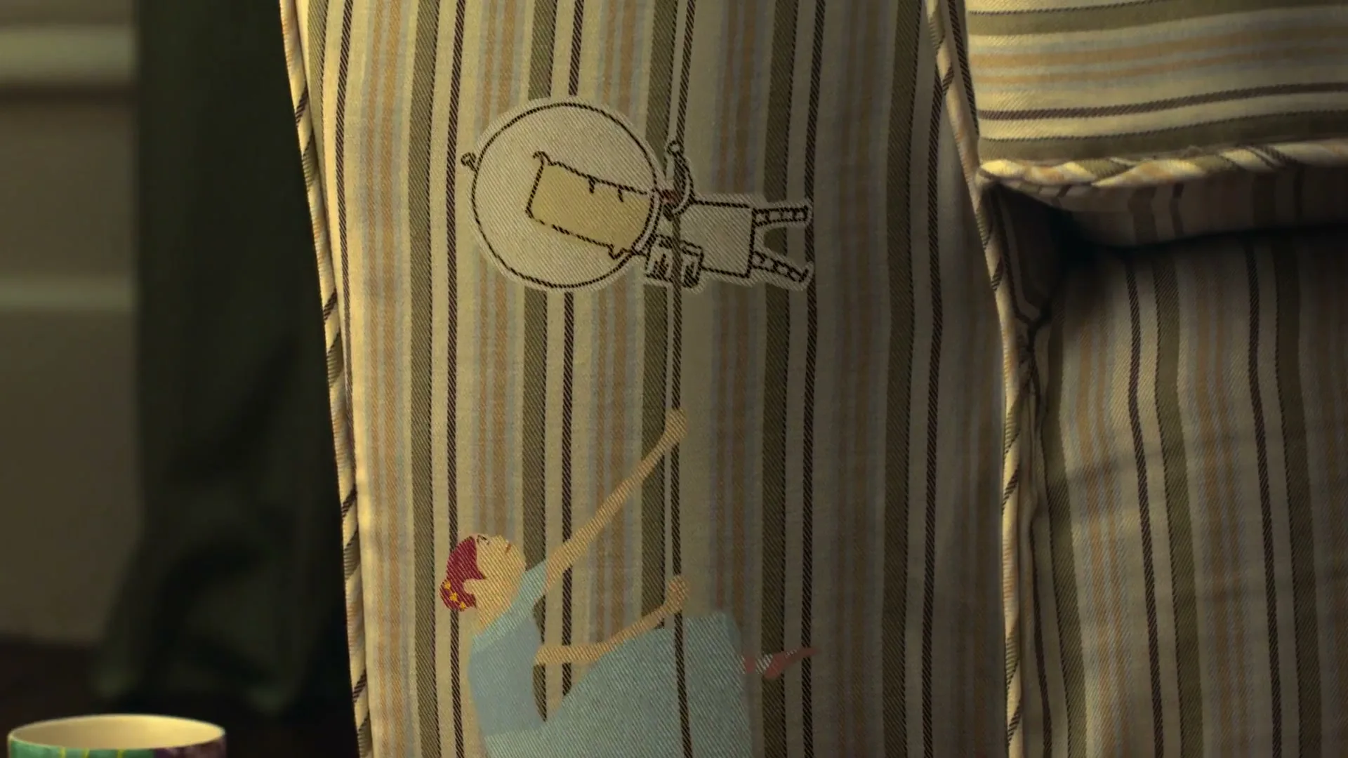 A closeup of a striped pillow with a cartoon drawing of a woman reaching for a man in a flying saucer, in a dimly lit room.