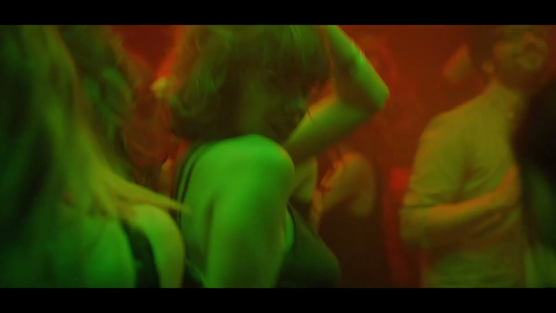 A diverse group of young adults dance closely together in a crowded, dimly lit nightclub, illuminated by green and orange party lights.