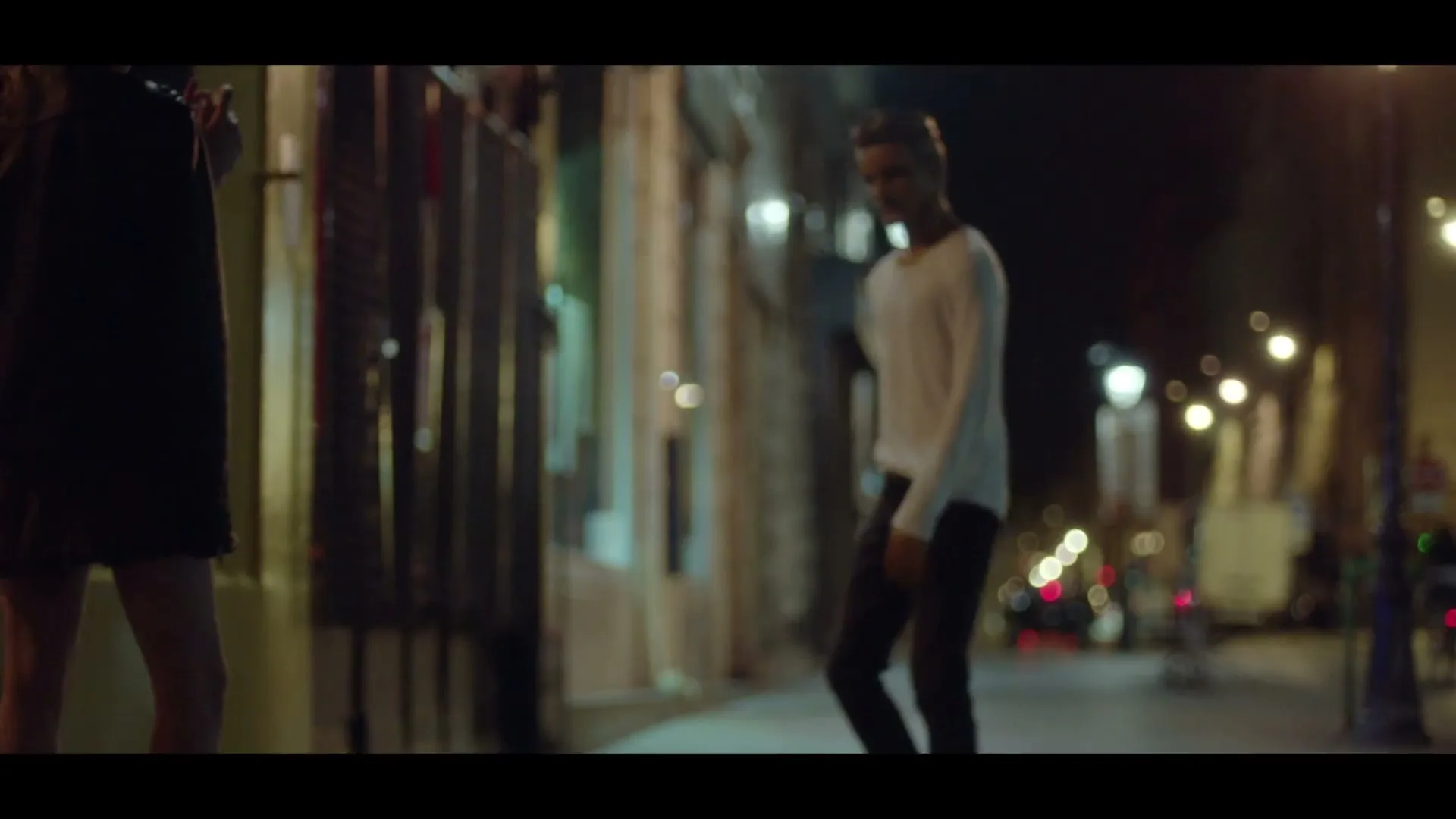 A young man in a white shirt and dark pants walks towards a young woman on a dark city street at night.