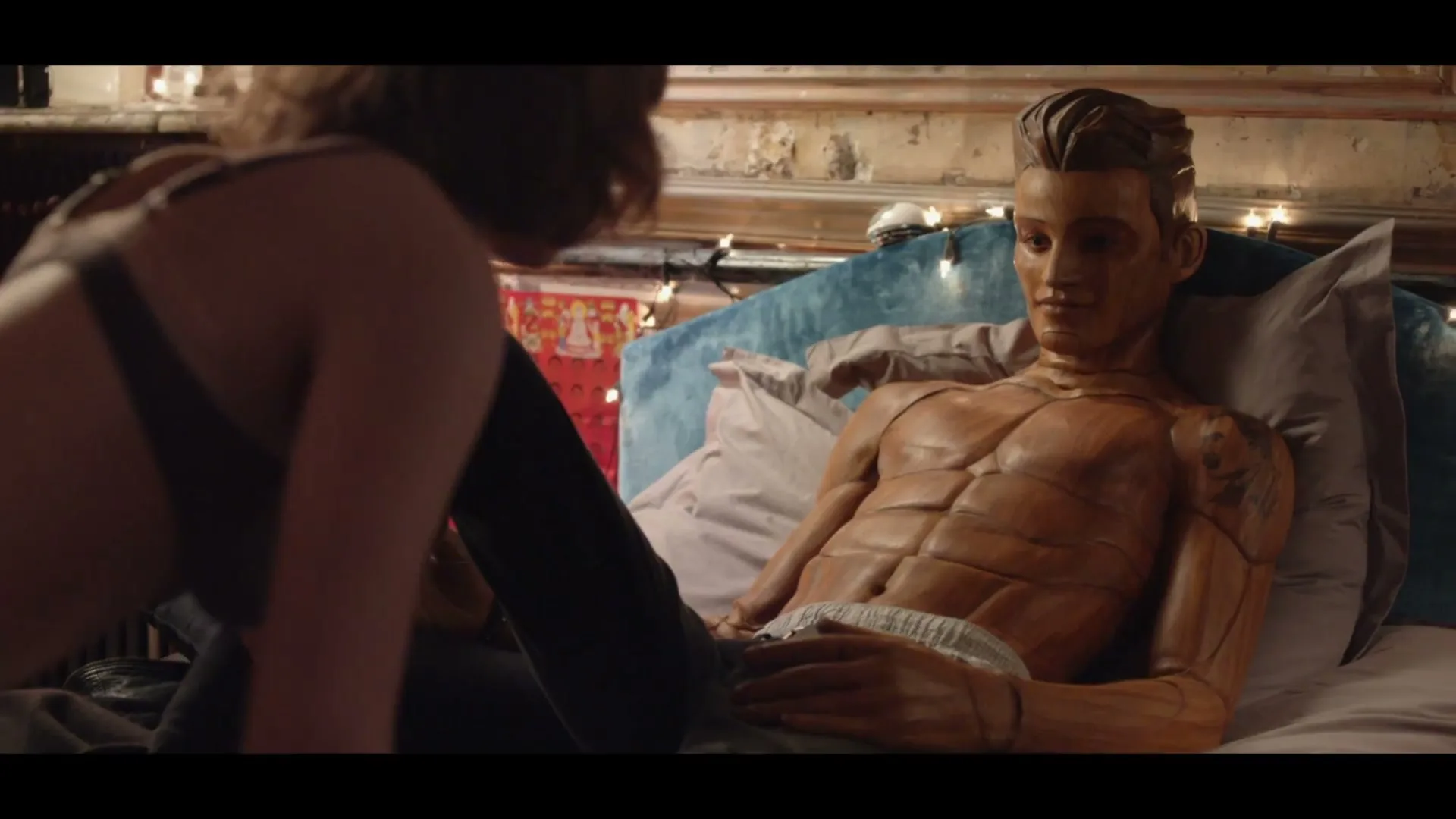 A woman in a bra leans over a wooden mannequin with sculpted abs, who is lying in bed in a bedroom.