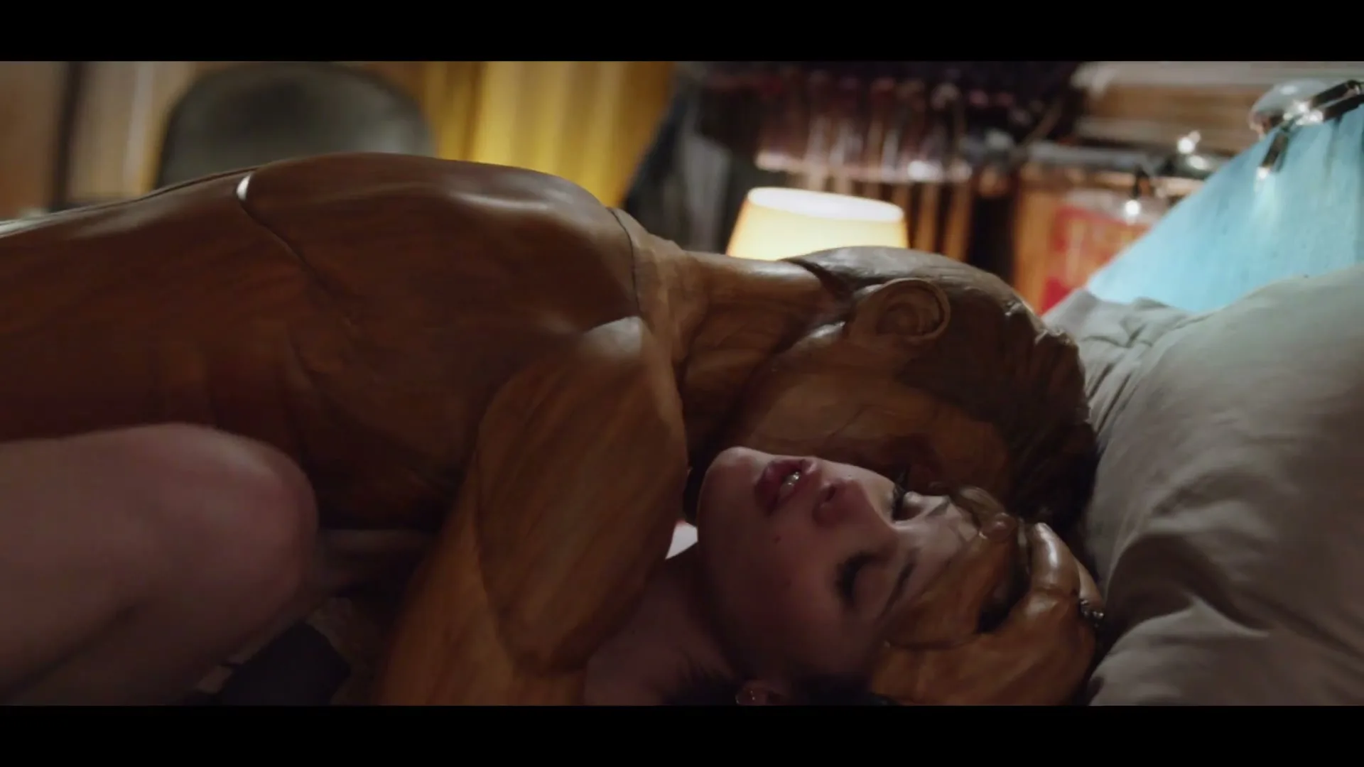 A young woman with light brown hair lies in bed, eyes closed, as a wooden mannequin-like figure leans over her.