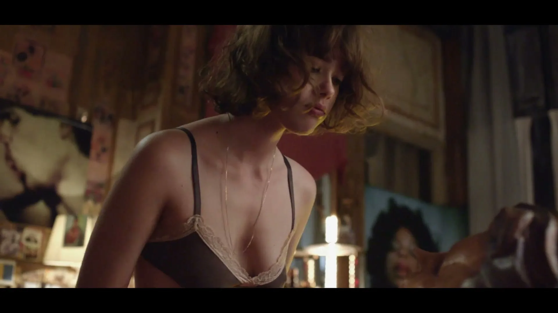 A young woman with short, curly hair and a bra looks down, seemingly in thought, in a dimly lit, cluttered bedroom.