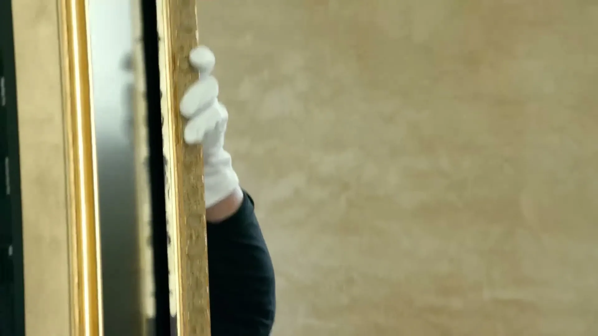 A person in white gloves carefully holds a large, ornate golden picture frame, partially visible against a light-colored wall.
