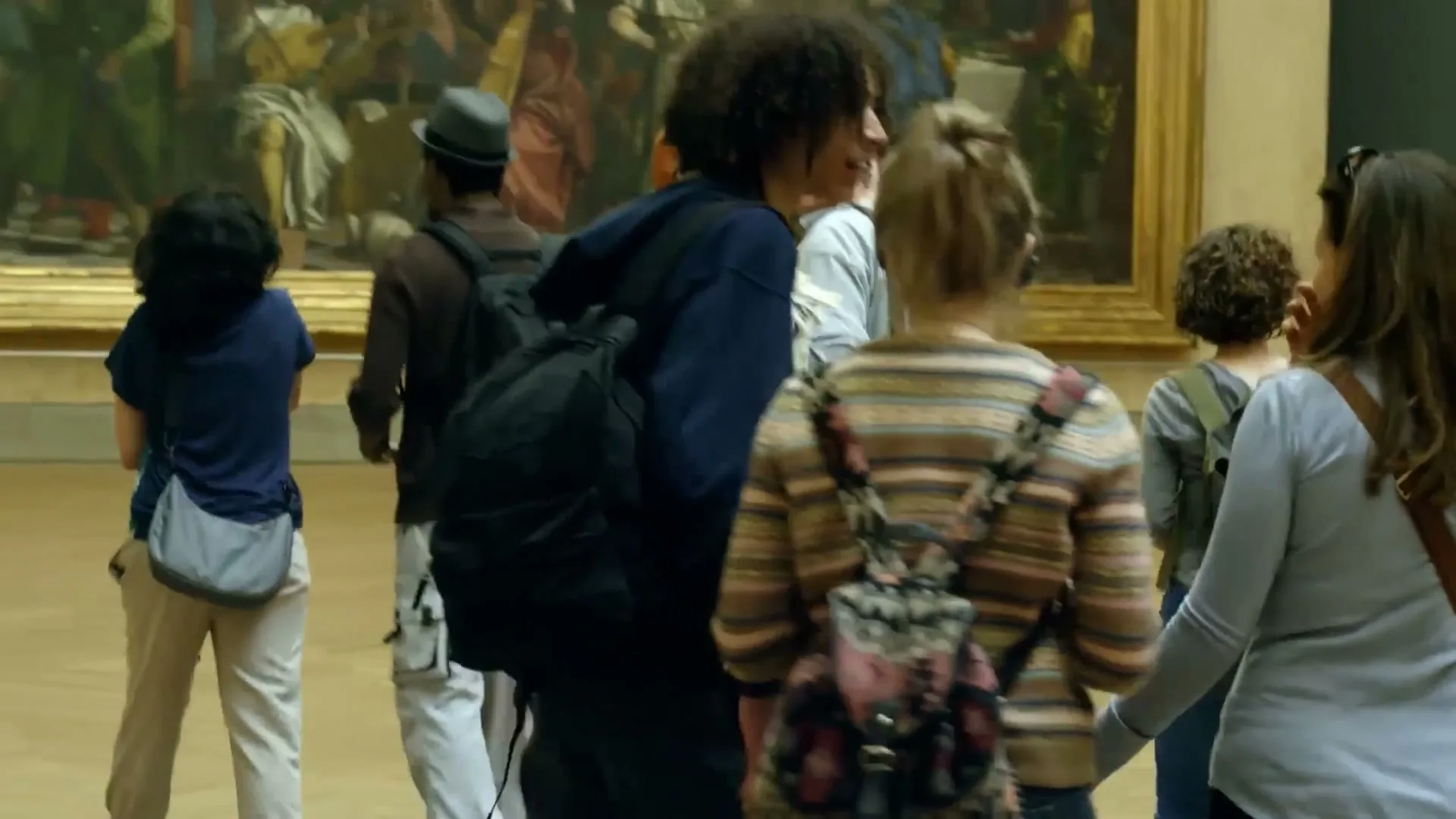 A diverse group of tourists, some with backpacks, walk through a museum gallery, looking at a large classical painting on the wall.