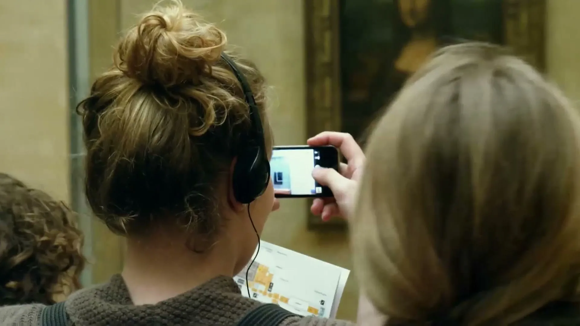 A young woman with headphones and a bun takes a photo of the Mona Lisa with her phone, while another person stands in front of her at the Louvre Museum.