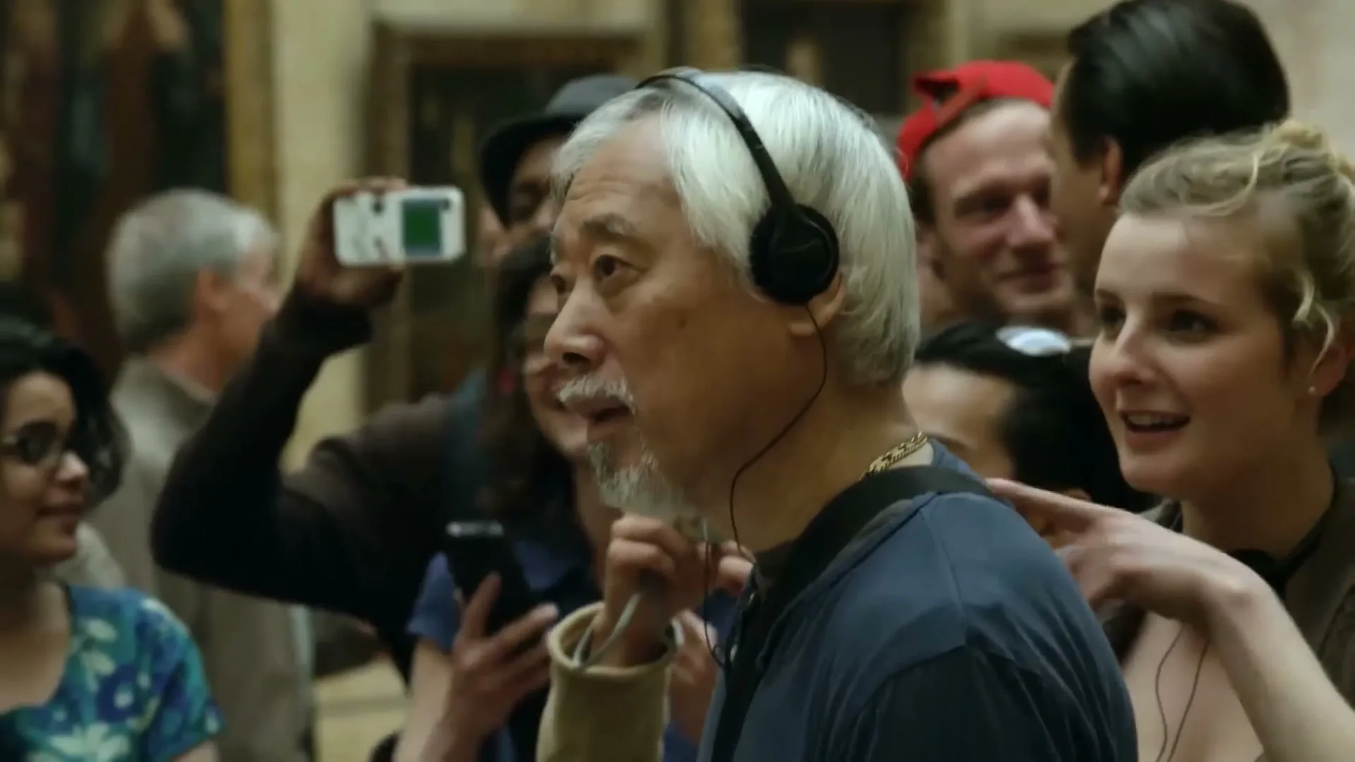 A diverse group of tourists, including an older Asian man with headphones, are looking at something off-screen in a museum.