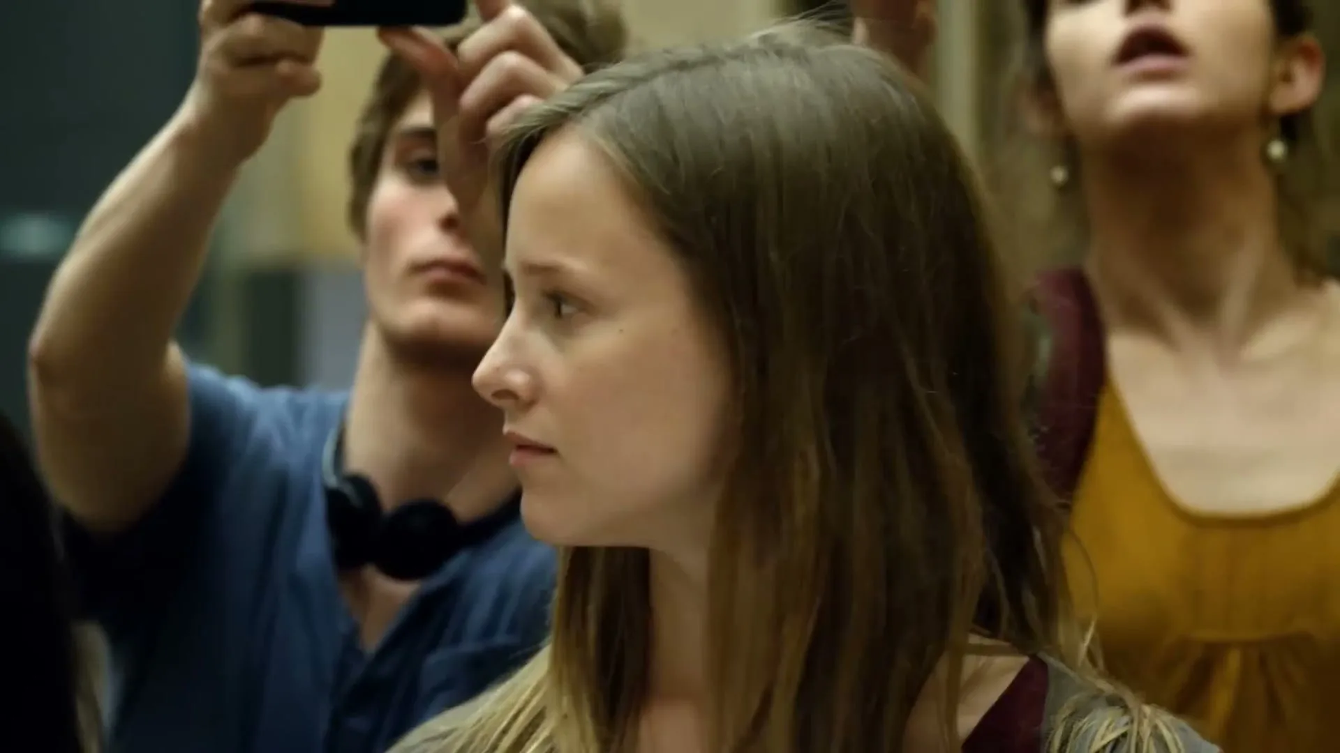 A young woman with long brown hair looks around, surrounded by other people taking photos with their phones in a museum.