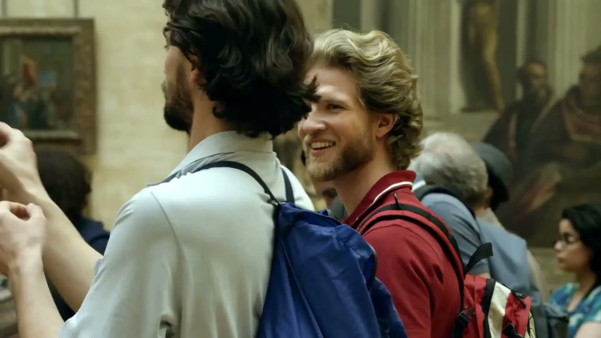 Two male tourists, one with a beard and dark hair, the other with blonde hair, smile while looking at art in a crowded museum.