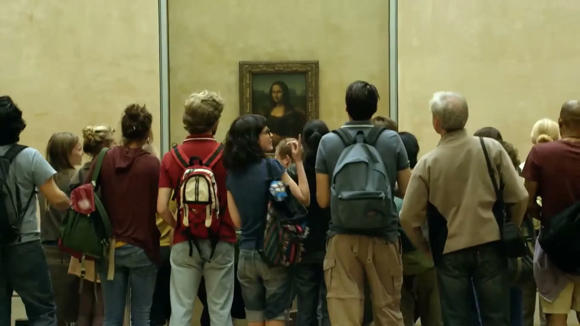 A diverse group of tourists, seen from behind, stand in a crowded museum gallery, looking at the Mona Lisa painting.
