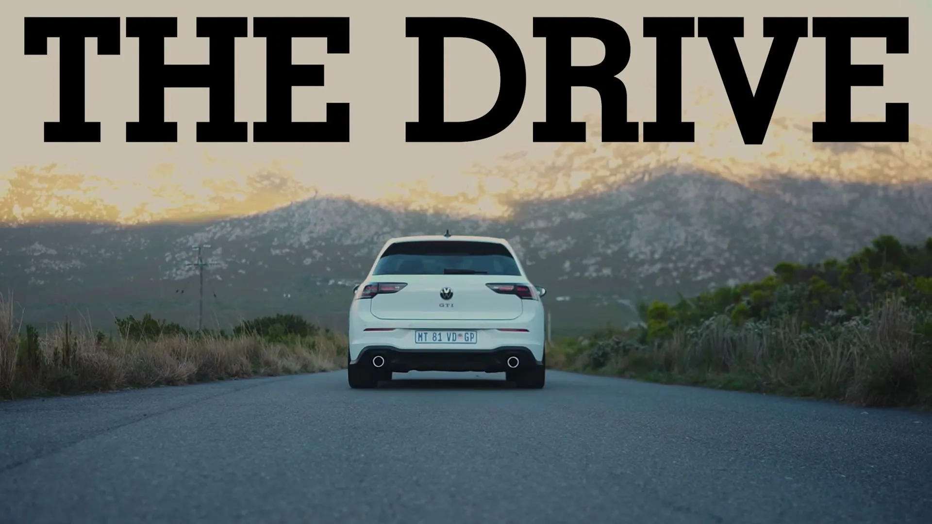A white Volkswagen Golf GTI drives away from the camera on a rural road, with mountains and a sunset in the background.