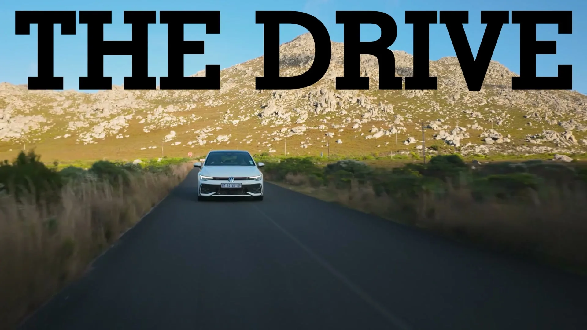 A white Volkswagen car drives towards the camera on a winding road through a mountainous, arid landscape under a clear blue sky, with "THE DRIVE" text overlaid.