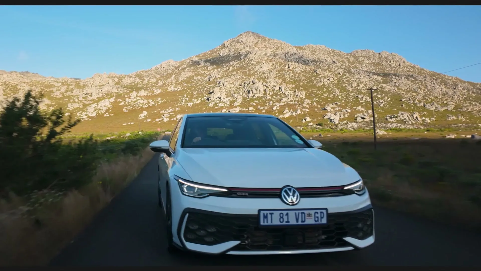 A white Volkswagen Golf GTI drives on a winding road through a mountainous landscape under a clear blue sky.