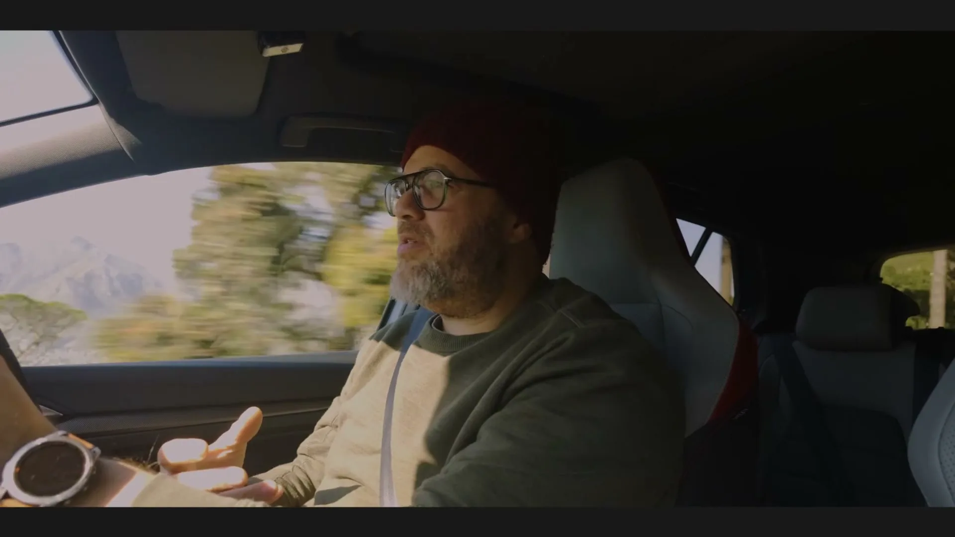A bearded man in a beanie and glasses drives a car, gesturing with his hands while talking, with blurred scenery passing by outside the window.