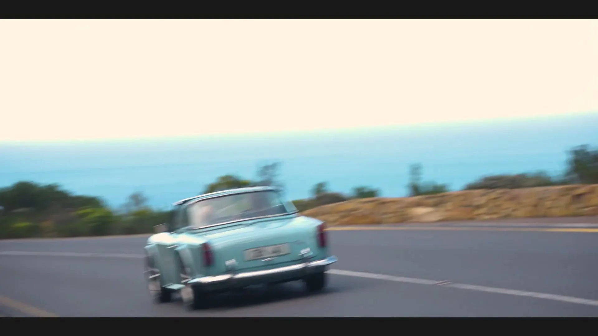 A vintage light blue convertible car drives along a coastal road, with a white SUV approaching from behind, under a bright sky.