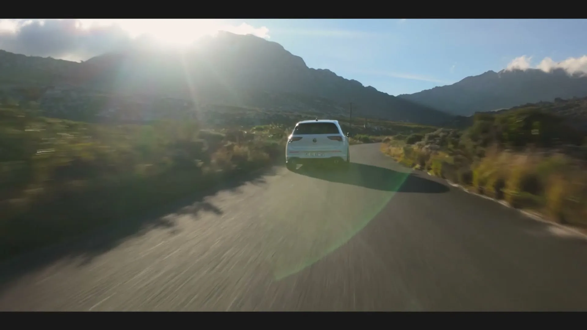 A white Volkswagen Golf GTI drives away from the camera on a winding road through a mountainous landscape under a bright, sunny sky.