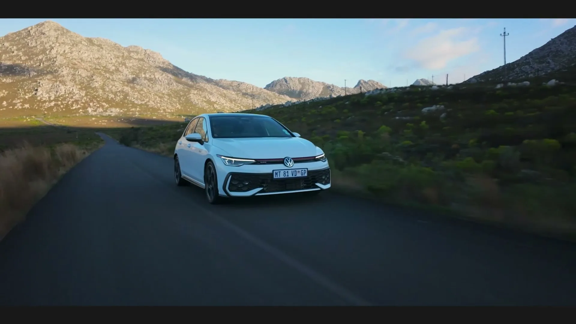 A white Volkswagen Golf GTI drives on a winding road through a mountainous landscape under a clear sky.