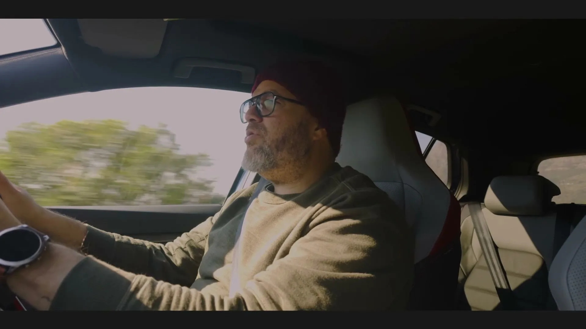A bearded man in a red beanie and glasses drives a car, looking intently at the road ahead on a sunny day.