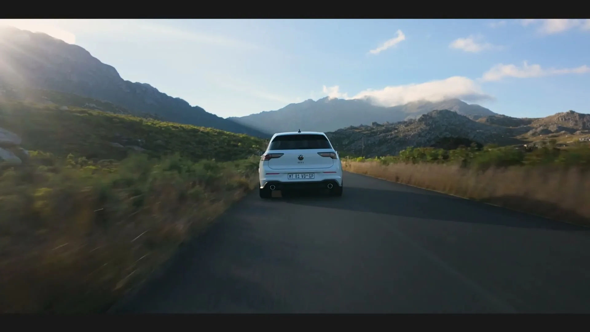 A white Volkswagen GTI drives away from the camera on a winding road through a mountainous landscape under a bright, sunny sky.