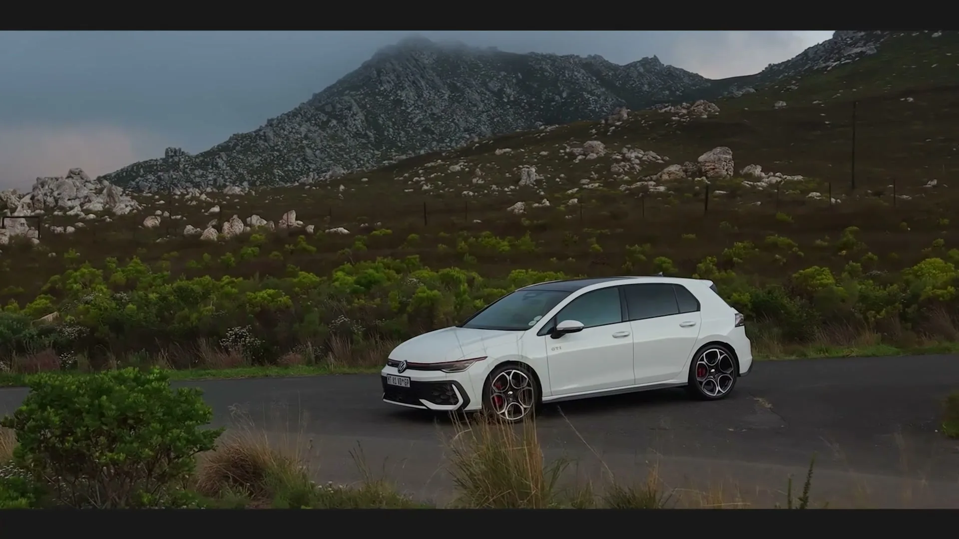 A white Volkswagen Golf GTI is parked on a winding road, surrounded by green bushes and rocky mountains under a cloudy sky.