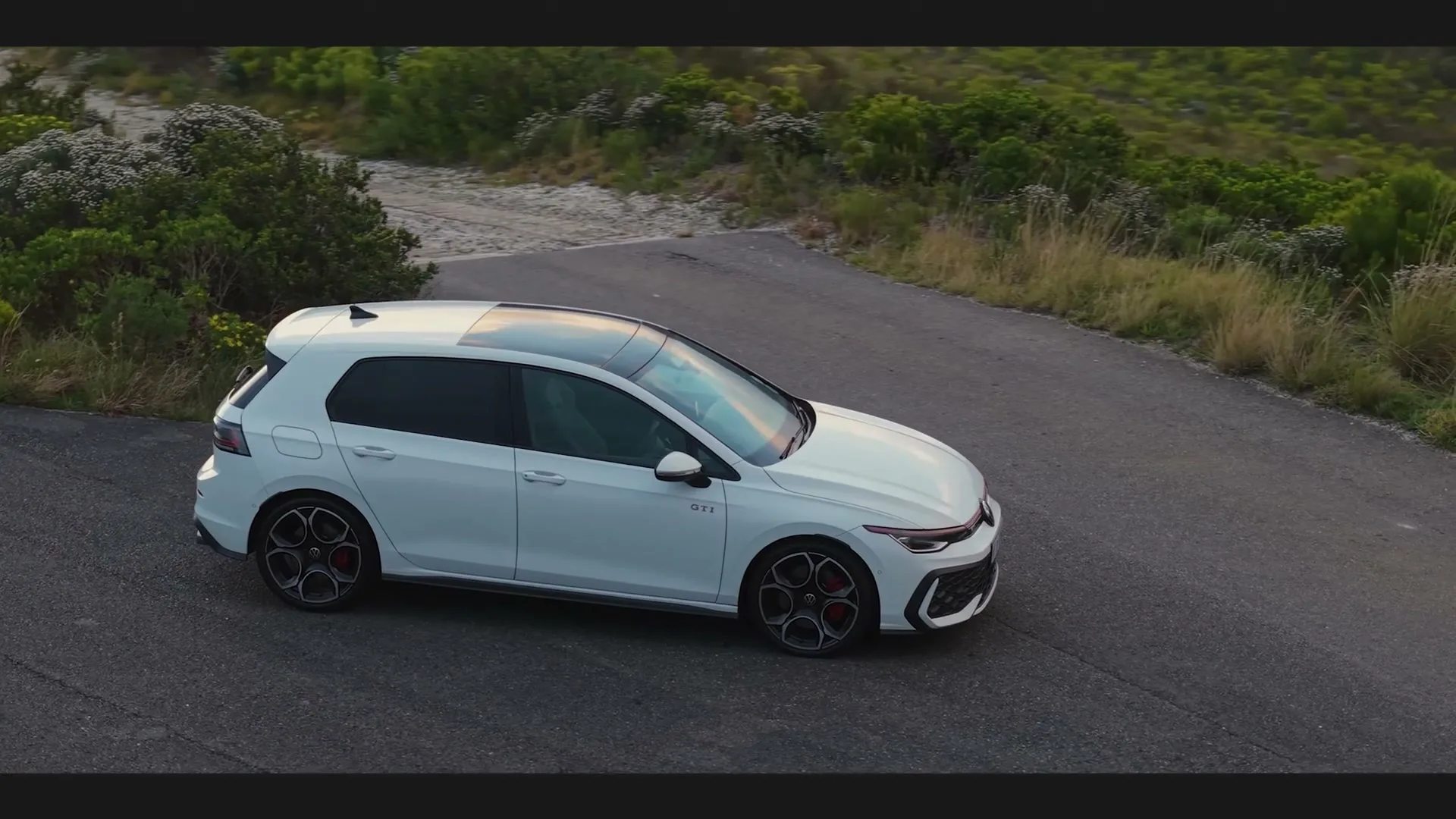 A white Volkswagen Golf GTI drives along a winding road surrounded by green bushes and a dirt path under a bright sky.