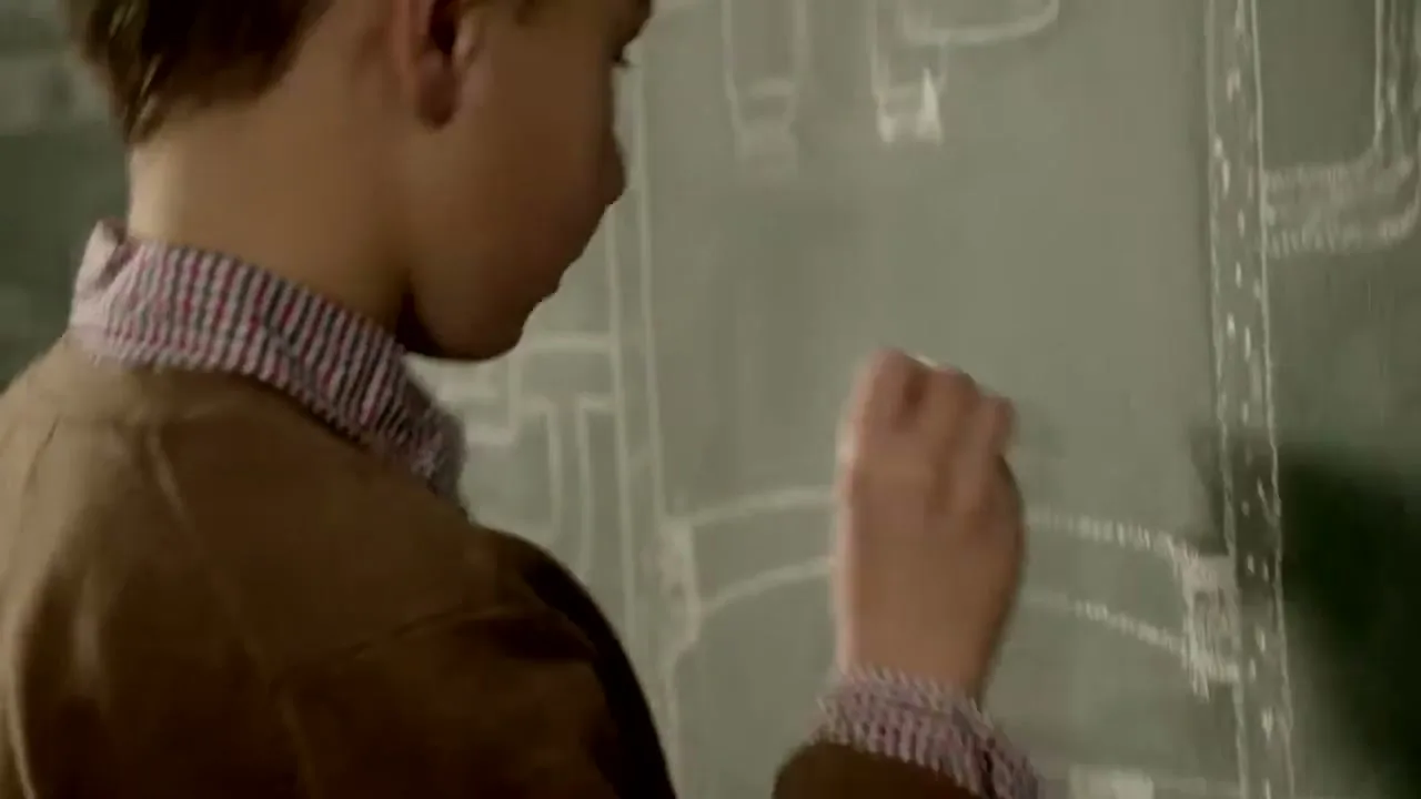 A young boy in a brown jacket and plaid shirt draws a complex plumbing diagram on a green chalkboard in a classroom.