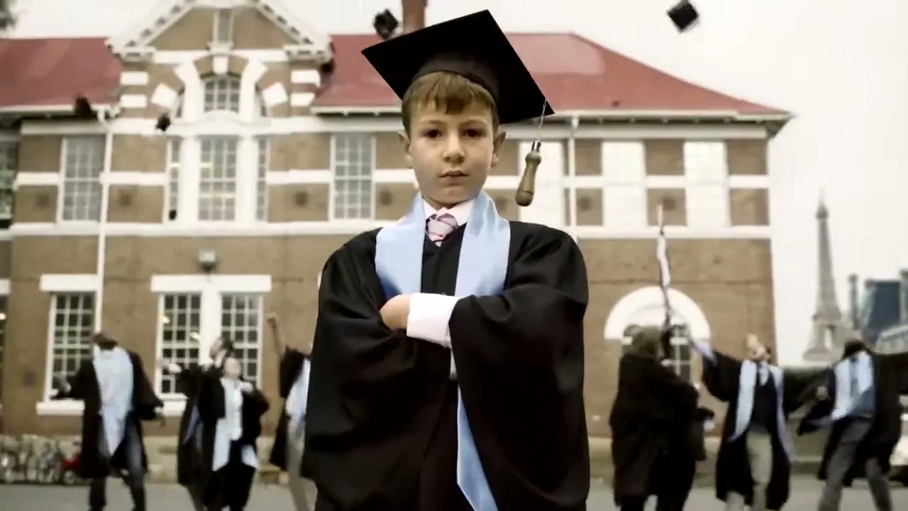 A young boy in a graduation cap and gown stands with arms crossed, looking directly at the camera, as other graduates celebrate in the background.