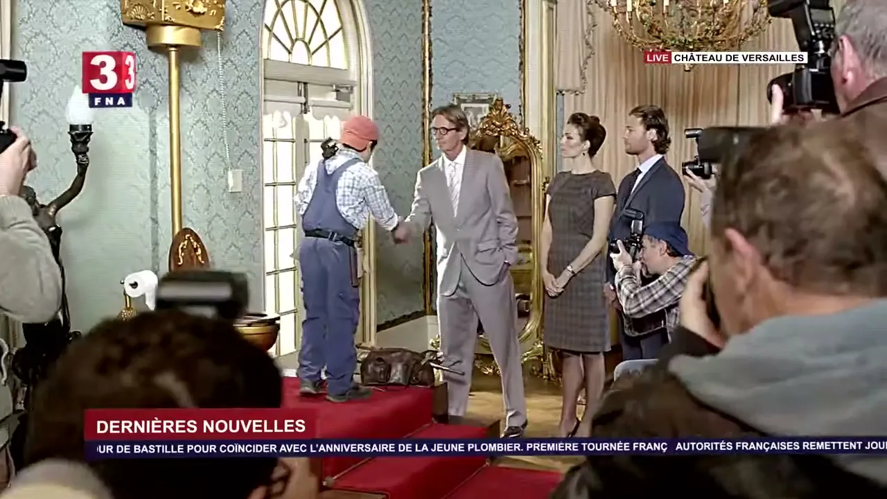 A young plumber in overalls shakes hands with a man in a suit, while a woman and another man stand by, surrounded by photographers in a lavish room.