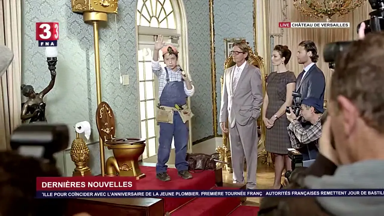 A young boy dressed as a plumber waves to a crowd of photographers, standing next to a golden toilet and a couple in a lavish room.
