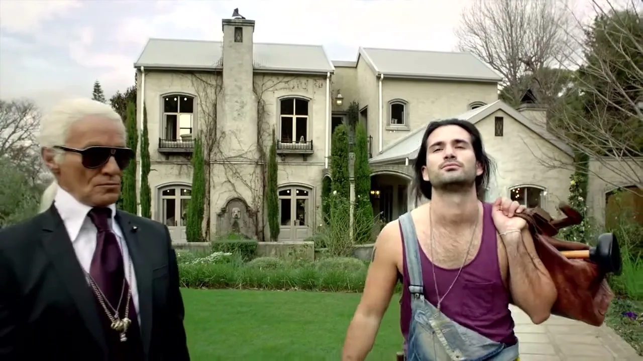 A young plumber with a tool bag walks past a man resembling Karl Lagerfeld in front of a large house.