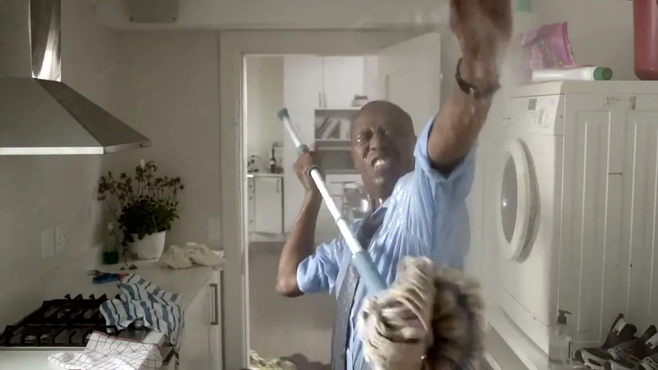 A man in a blue shirt and tie is drenched in water, holding a mop, looking distressed in a kitchen with a burst pipe.