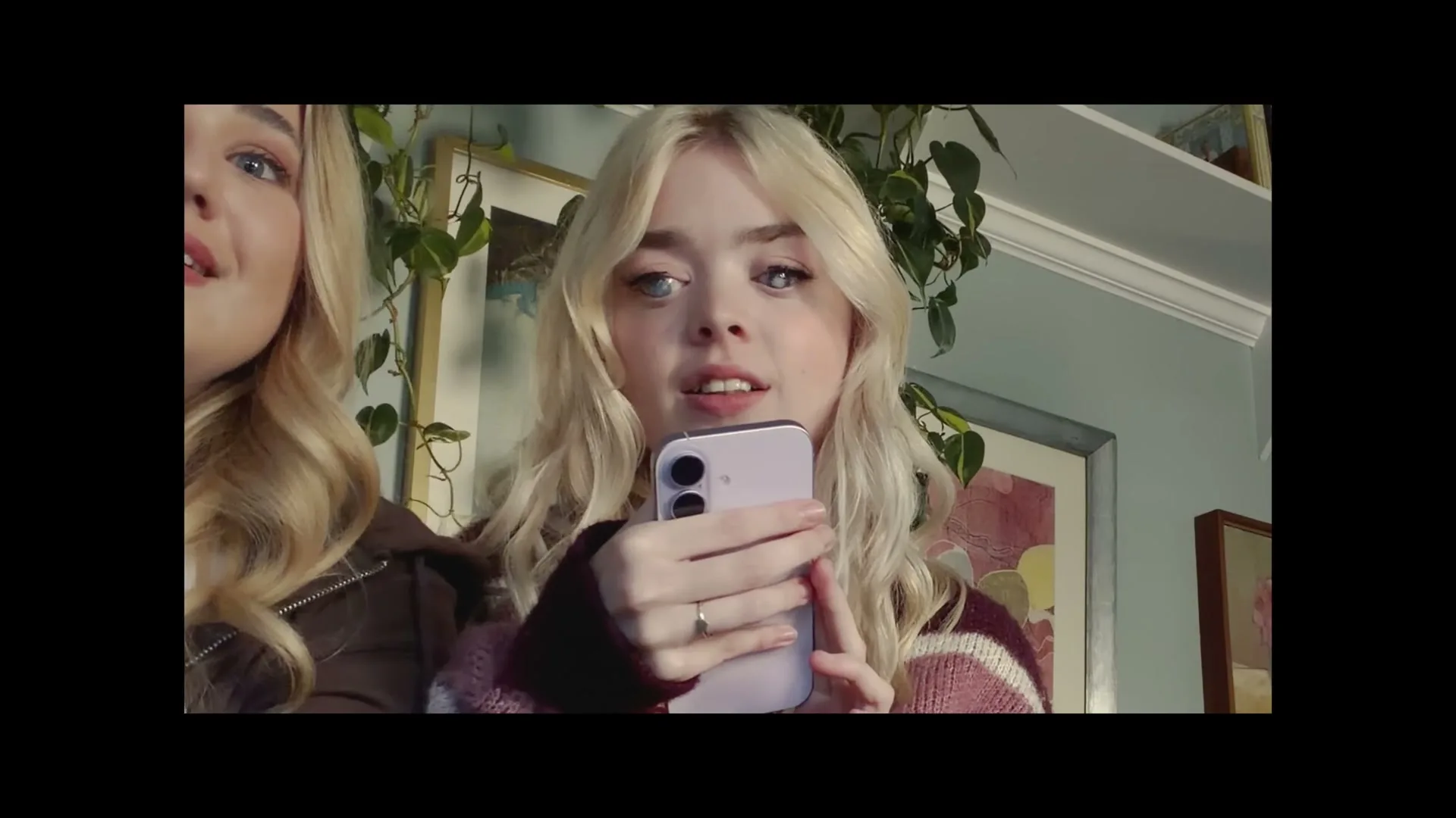 Two young women with blonde hair are looking at a phone, one holding it, as they react with surprise and excitement in a cozy living room.