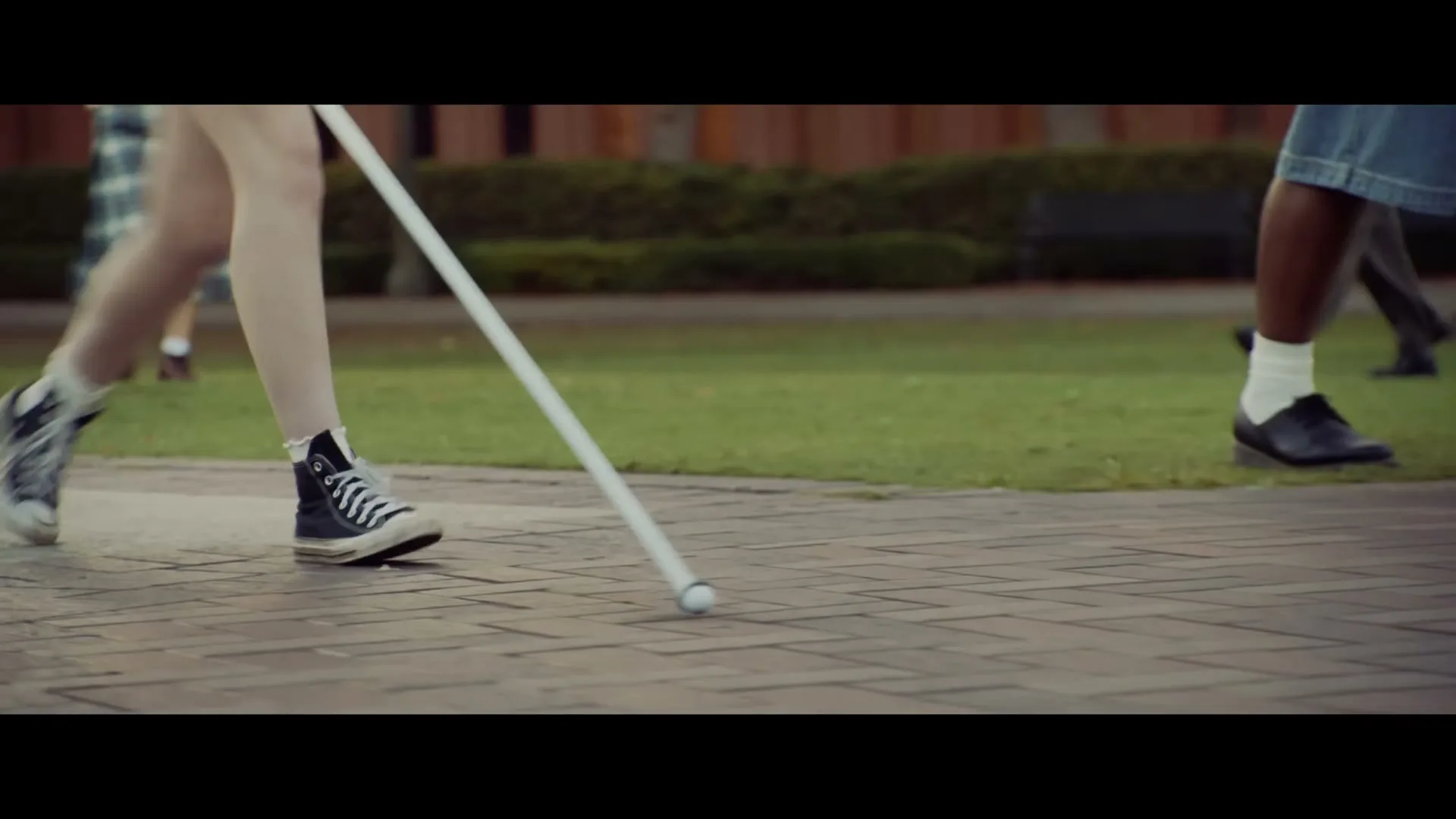 A young person with a white cane walks on a paved path, their legs and feet visible, with other people walking in the background on a college campus.