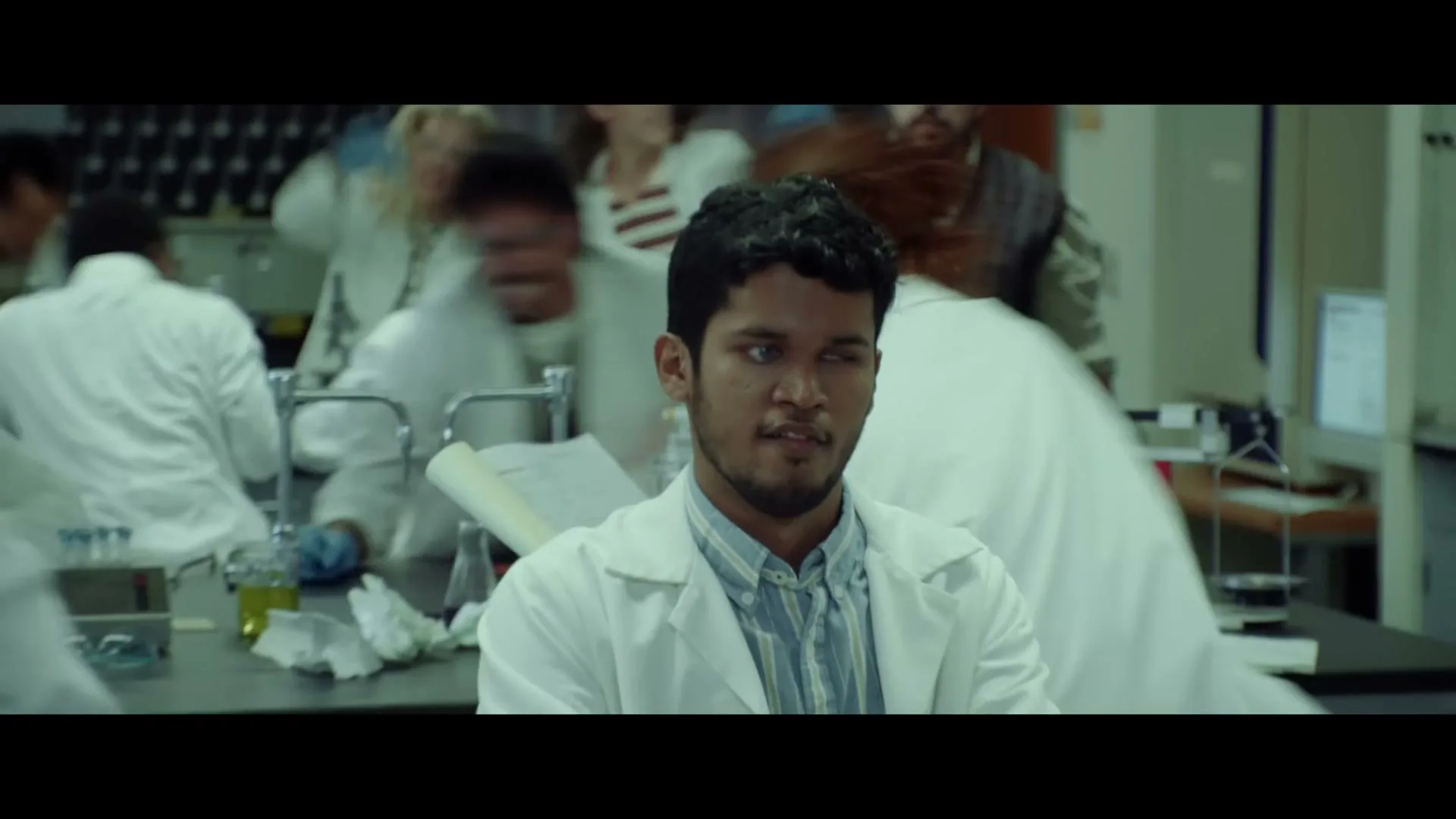 A young man in a lab coat sits at a desk, looking around as other scientists in lab coats move quickly around him in a laboratory.