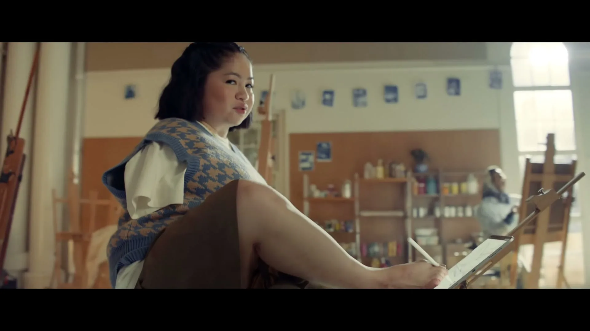 A young Asian woman with short dark hair, wearing a patterned vest and white shirt, sits cross-legged, drawing on a tablet with her foot in an art studio.
