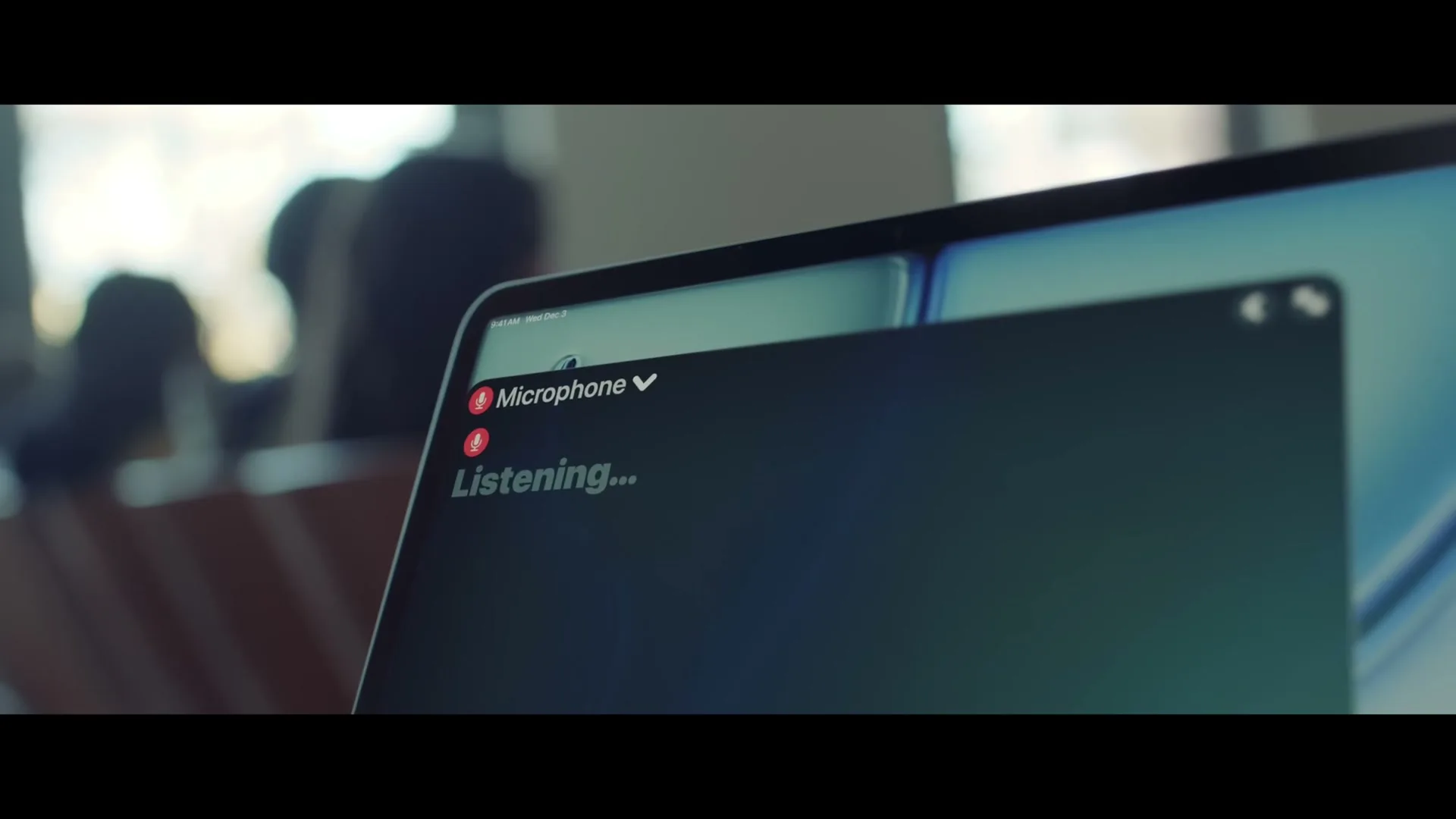 A close-up shot of a laptop screen showing a "Microphone Paused..." then "Listening..." interface, with a blurred background of an interior space.