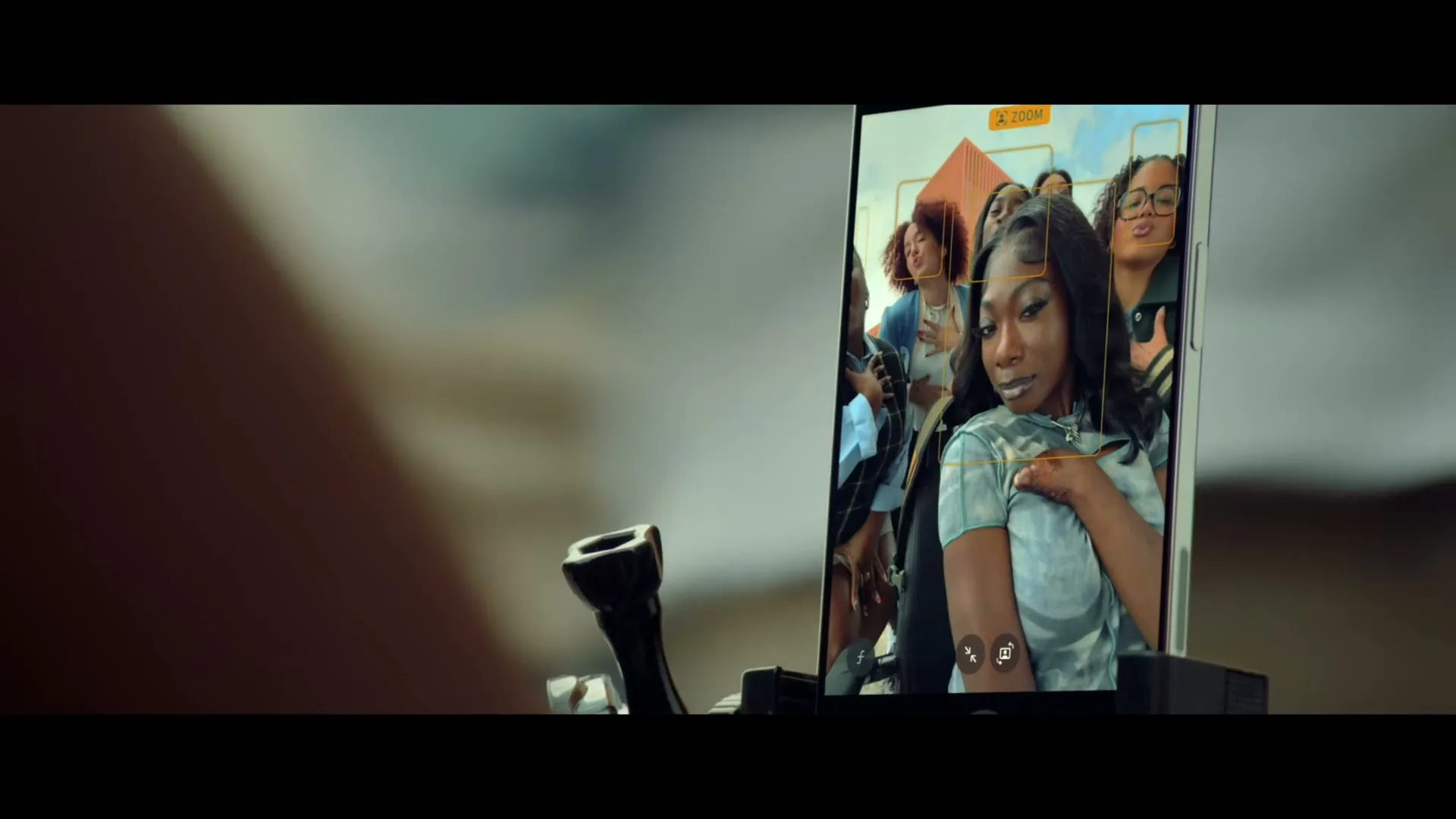A close-up shot shows a smartphone screen displaying a group of young women posing for a selfie, held by an unseen person outdoors.