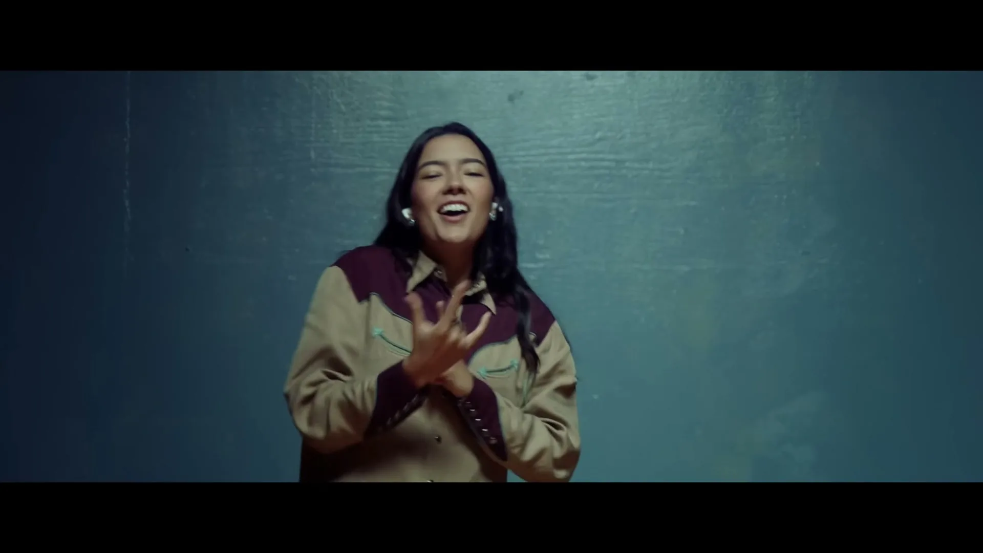 A young woman with long dark hair, wearing a beige and maroon western shirt, dances and gestures with her hands in front of a dark blue wall.