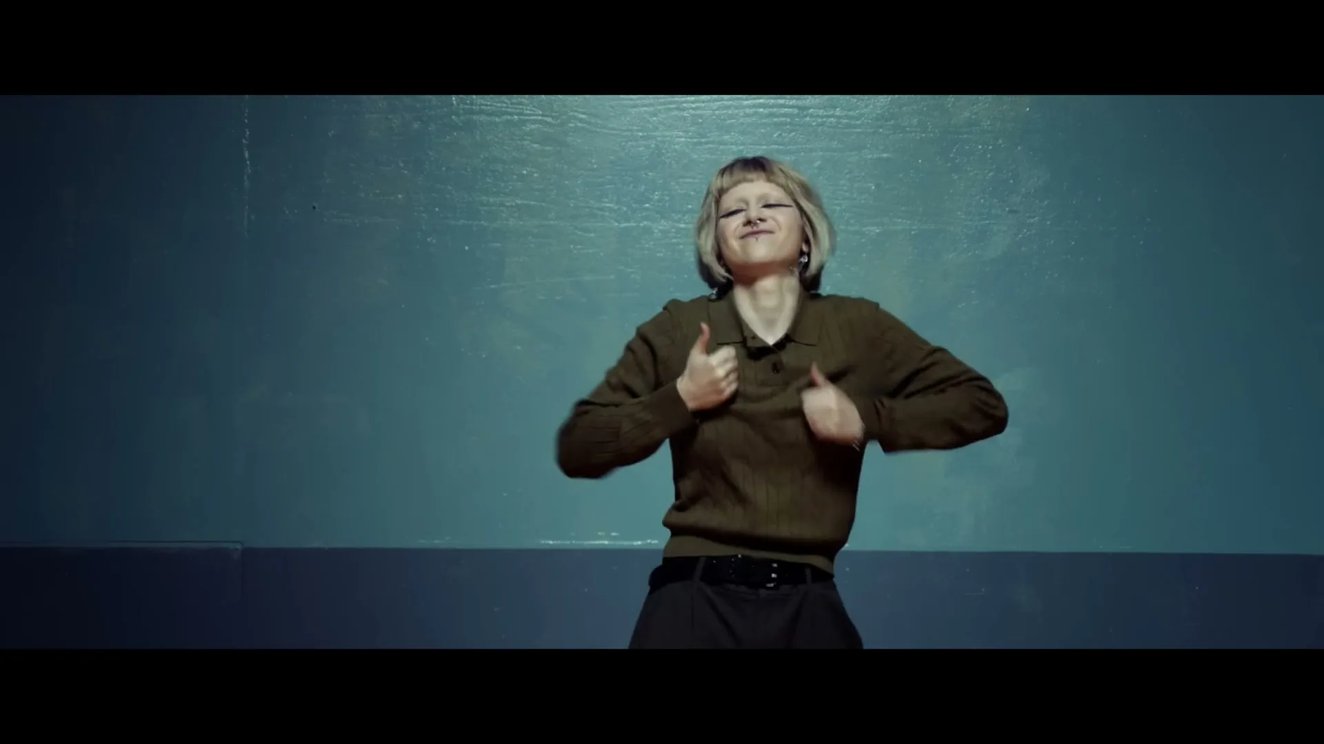 A young woman with short blonde hair and dark eye makeup dances energetically in a studio with a blue wall.