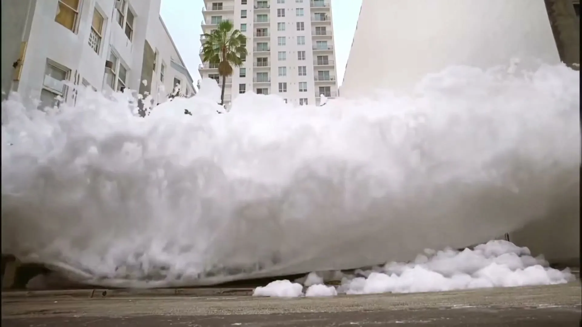 A large wave of white foam surges forward, filling the street between buildings, with a palm tree and tall building visible in the background.