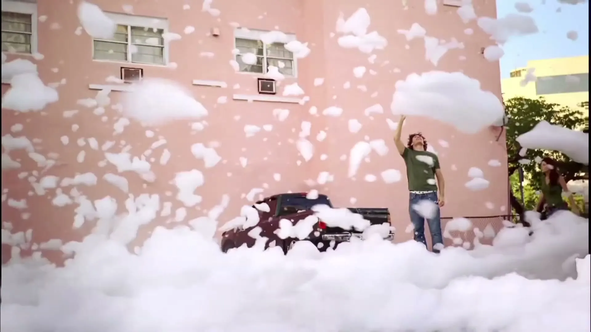 A young man in a green shirt and jeans stands in a street covered in foam, reaching up to catch a foam cloud, with a pink building and a truck in the background.