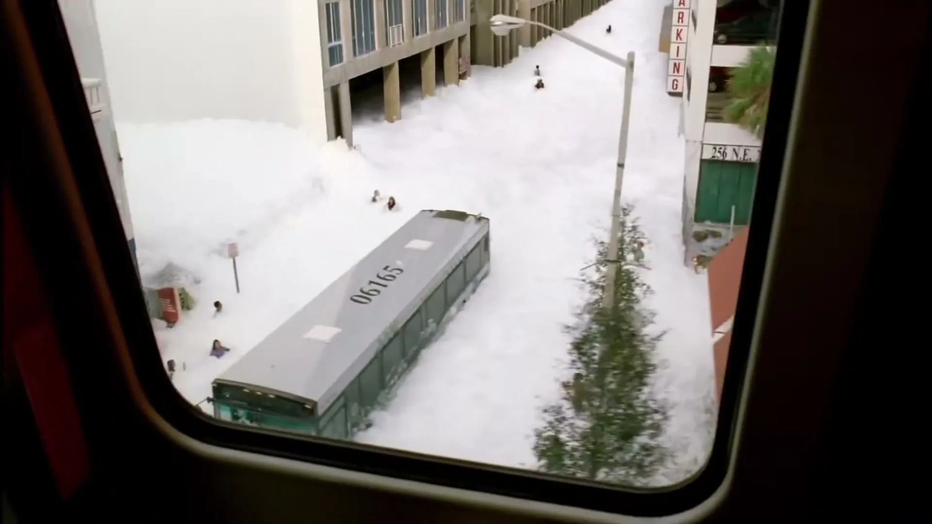 A high-angle shot from a moving vehicle shows a city street covered in foam, with a bus and people partially submerged.