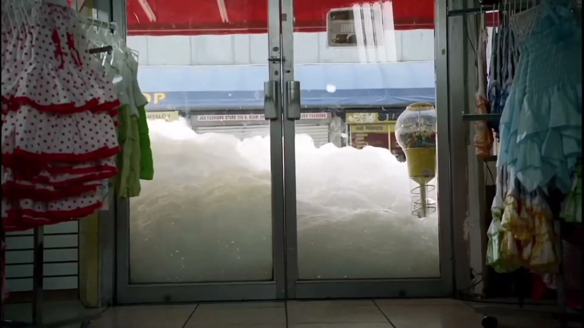 A wide shot from inside a clothing store shows the street outside filled with white foam, almost reaching the top of the glass doors.